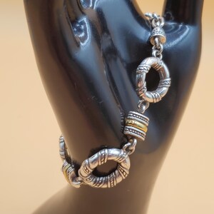 Op de afbeelding: Zilverkleurige armband met decoratieve cirkelvormige schakels en goudkleurige accenten. De armband wordt getoond op een zwart handvormig display tegen een lichtoranje achtergrond. De armband heeft een textuur.