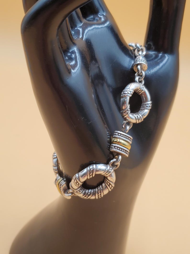 Op de afbeelding: Zilverkleurige armband met decoratieve cirkelvormige schakels en goudkleurige accenten. De armband wordt getoond op een zwart handvormig display tegen een lichtoranje achtergrond. De armband heeft een textuur.