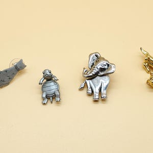 Puede incluir: Cuatro broches con temática de animales. Un broche de gato plateado, un broche de tortuga plateado, un broche de elefante plateado y un broche de perro dorado. Los broches están sobre una superficie de color beige claro.