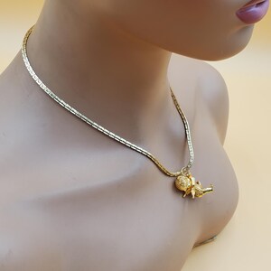Pu&ograve; includere: Una collana dorata con una catena delicata e un ciondolo. Il ciondolo presenta una piccola figura dettagliata. La collana &egrave; esposta su un manichino con uno sfondo neutro.
