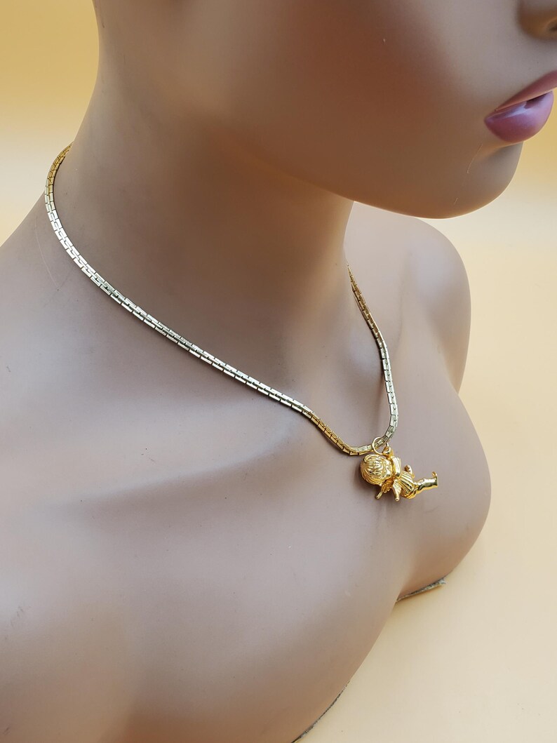 Pu&ograve; includere: Una collana dorata con una catena delicata e un ciondolo. Il ciondolo presenta una piccola figura dettagliata. La collana &egrave; esposta su un manichino con uno sfondo neutro.