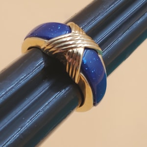 May include: A gold-toned ring with a deep blue, speckled enamel band. The ring features a crisscross design in gold, creating a striking contrast with the blue enamel. The ring is a piece of jewelry.