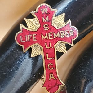 May include: A red cross pin with gold accents. The pin reads "W.M.S.U.L.C.A. LIFE MEMBER".