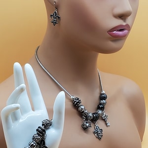 Puede incluir: Un conjunto de joyas plateadas con collar, pulsera y pendientes. El collar y la pulsera tienen cuentas negras y plateadas con dijes de flor de lis, corona y llave. Los pendientes tienen forma de flor de lis.
