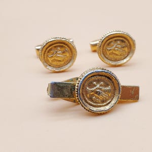 May include: A set of gold-toned metal accessories including two round cufflinks and one tie bar. Each piece features a circular emblem with a raised design of two hands shaking, surrounded by a beaded border and a smooth metallic edge.