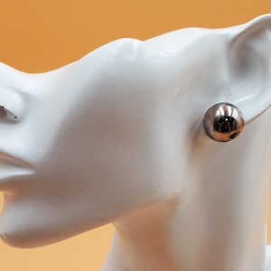 May include: A single silver-colored ball earring is displayed on a white mannequin head. The earring is round and shiny, reflecting light. The background is a solid orange color.