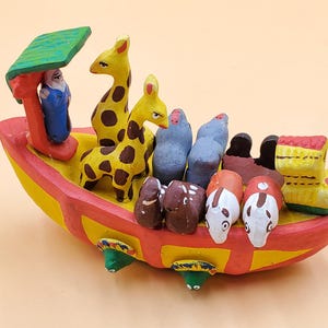 May include: A brightly painted wooden Noah's Ark toy. The ark is red, yellow, and green, with animal figures such as giraffes and birds. A figure of a person is under a green roof. The toy is designed for children and is a traditional toy.
