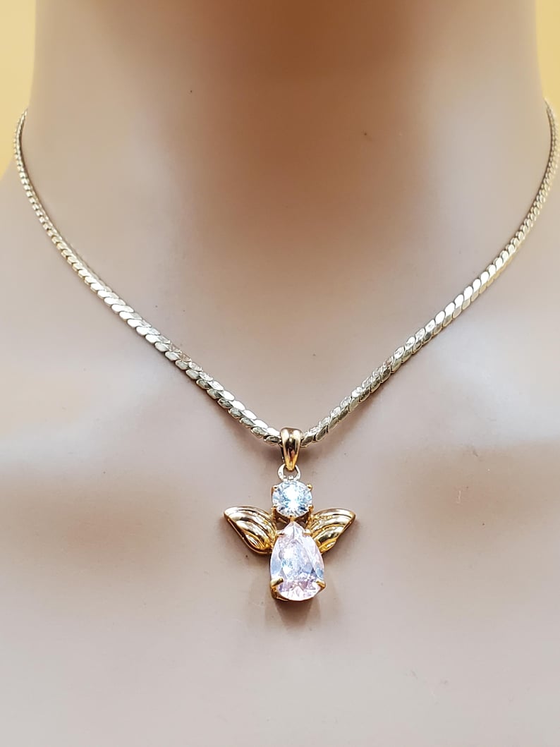 May include: A gold-toned necklace featuring an angel pendant. The pendant has a pear-shaped, clear gemstone and a smaller, round, clear gemstone. The angel has gold-colored wings. The necklace chain is a flat, linked design.