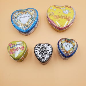 May include: Five heart-shaped decorative tins. The tins feature various designs, including floral patterns, the word "Brighton", and the word "Love". Colors include blue, yellow, green, black, and white. The tins are likely used for small items or as decorative pieces.