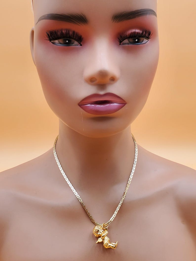 Pu&ograve; includere: Una collana color oro con una catena a maglie rettangolari e un ciondolo raffigurante un piccolo cherubino. Il cherubino ha le ali ed &egrave; seduto. La collana &egrave; esposta su un manichino.