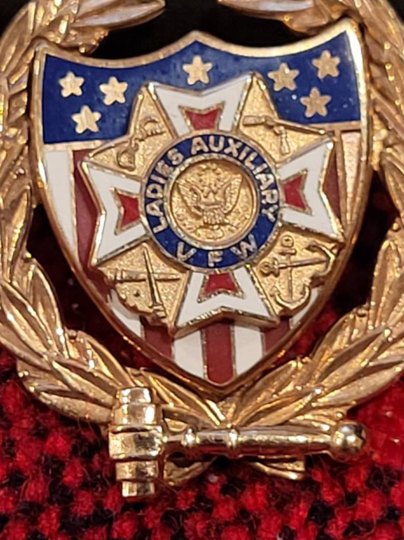 Puede incluir: Un pin de tono dorado con un dise&ntilde;o en forma de escudo. El escudo presenta una bandera estadounidense azul y blanca con estrellas, una cruz roja y blanca y un &aacute;guila dorada. El texto "LADIES AUXILIARY" est&aacute; escrito alrededor del &aacute;guila. El escudo est&aacute; rodeado por una corona de hojas doradas.