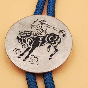 May include: A silver-toned bolo tie featuring a black graphic of a cowboy riding a bucking bronco. The round pendant is connected to a blue braided cord.