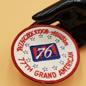 May include: White embroidered patch with red trim, featuring the text "Winchester-Yadkin 77th Grand American" and a blue and red rectangle with the number "76" inside. The patch is surrounded by white stars.