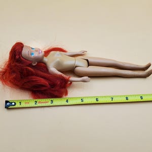 May include: A red-haired doll with blue eyes and a nude body. The doll is lying on a beige surface with a yellow measuring tape.