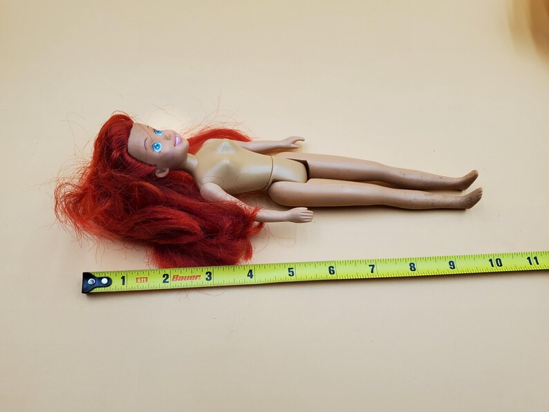 May include: A red-haired doll with blue eyes and a nude body. The doll is lying on a beige surface with a yellow measuring tape.