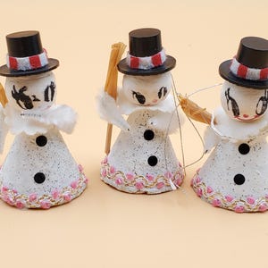Puede incluir: Tres figuras de muñecos de nieve vintage. Cada muñeco de nieve tiene un sombrero de copa negro con una banda a rayas rojas y blancas, un cuerpo blanco con detalles de botones negros y un ribete rosa y dorado en la base. Uno sostiene una escoba.