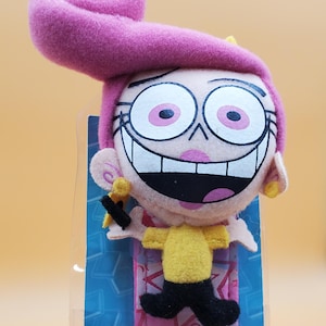 May include: A plush doll of a cartoon character with a large pink hair swirl, large eyes, and a wide smile. The doll wears a yellow top and black pants. The Nickelodeon logo is visible at the bottom.