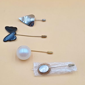 May include: Four decorative pins on a light orange background. The pins include a silver leaf shape, a black butterfly, a white pearl, and an oval-shaped pin with a gold-coloured frame and a white centre.