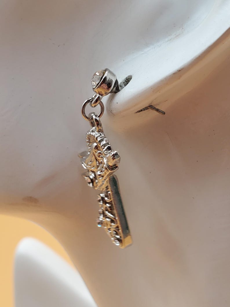K&ouml;nnte beinhalten: Silberfarbener Ohrring mit dekorativem Design. Der Ohrring hat einen kleinen, klaren Edelstein oben und einen verzierten, rechteckigen Anh&auml;nger. Das Design umfasst Schn&ouml;rkel und andere dekorative Elemente. Der Ohrring wird vor wei&szlig;em Hintergrund gezeigt.