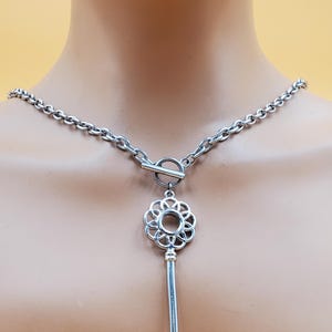 May include: Silver-toned necklace featuring a key pendant. The key has a decorative floral design at the top and a classic key shape. The necklace has a chain with a toggle clasp.