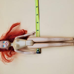 May include: A red-haired doll with blue eyes and a nude body. The doll is lying on its back with its legs extended. A measuring tape is visible in the background.
