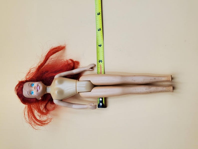 May include: A red-haired doll with blue eyes and a nude body. The doll is lying on its back with its legs extended. A measuring tape is visible in the background.
