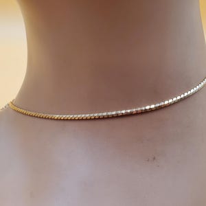 May include: A gold-coloured necklace with a delicate, textured chain. The necklace is shown on a mannequin, highlighting its design and the way it lays against the skin. The clasp is visible at the back. A simple, elegant piece of jewellery.