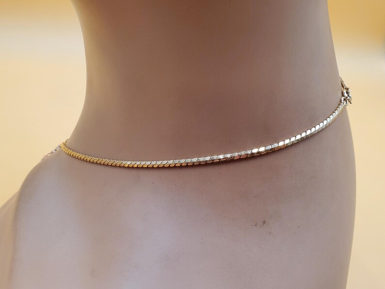 May include: A gold-coloured necklace with a delicate, textured chain. The necklace is shown on a mannequin, highlighting its design and the way it lays against the skin. The clasp is visible at the back. A simple, elegant piece of jewellery.