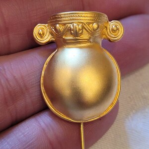 May include: A gold-colored metal pin in the shape of a vase with a decorative top and a round body. The pin has a long, thin stem.