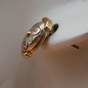 May include: A two-tone ring with gold and silver tones. The silver portion features an engraved swirl and diamond pattern. The ring has a gold accent on the top and bottom. The ring is set against a white background.