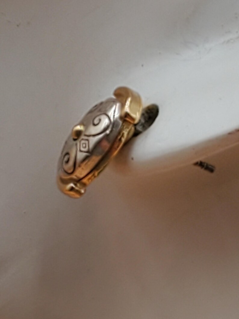 May include: A two-tone ring with gold and silver tones. The silver portion features an engraved swirl and diamond pattern. The ring has a gold accent on the top and bottom. The ring is set against a white background.