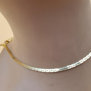 Pu&ograve; includere: Una collana dorata con un design a maglie piatte e rettangolari. La collana &egrave; esposta su un manichino e ha una chiusura a fermaglio. Il gioiello &egrave; uno stile classico, adatto a varie occasioni.