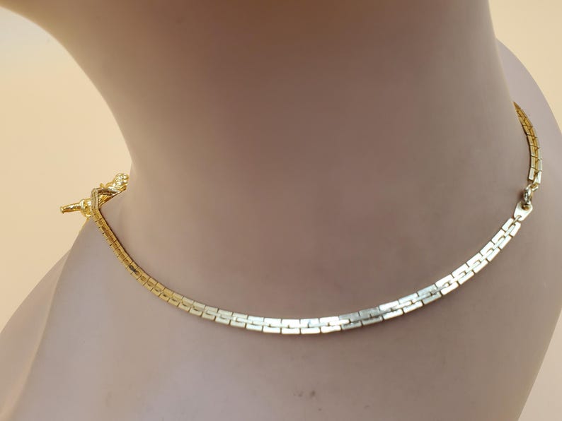 Pu&ograve; includere: Una collana dorata con un design a maglie piatte e rettangolari. La collana &egrave; esposta su un manichino e ha una chiusura a fermaglio. Il gioiello &egrave; uno stile classico, adatto a varie occasioni.