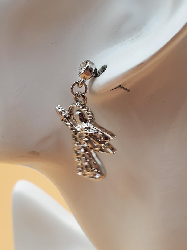 K&ouml;nnte beinhalten: Ein silberner Ohrring mit einem kleinen, detaillierten Engelanh&auml;nger. Der Ohrring ist an einem wei&szlig;en Ohr befestigt. Der Engelanh&auml;nger hat strukturierte Fl&uuml;gel und einen detaillierten K&ouml;rper. Der Ohrring ist f&uuml;r das Tragen in einem durchbohrten Ohr konzipiert.