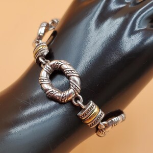 Op de afbeelding: Zilverkleurige armband met gouden accenten. De armband heeft een gestructureerd, cirkelvormig middelpunt en een knevelsluiting. Het ontwerp omvat gedraaide touwdetails en goudkleurige banden. De armband wordt op een zwart oppervlak gepresenteerd.