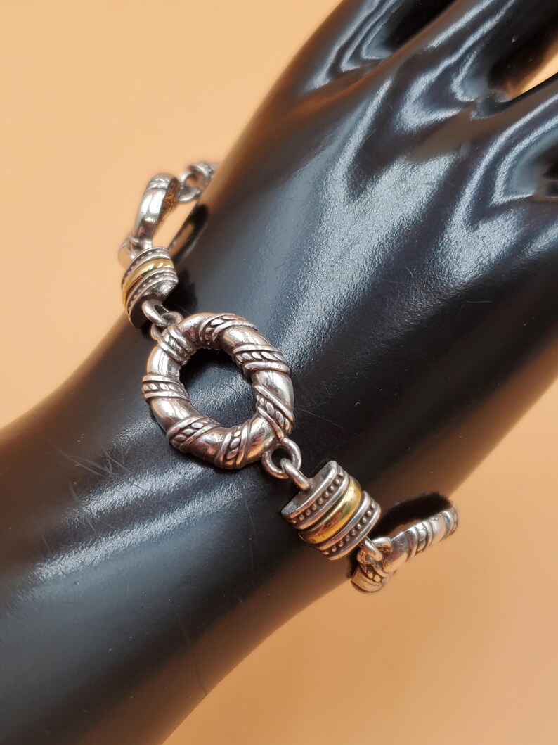 Op de afbeelding: Zilverkleurige armband met gouden accenten. De armband heeft een gestructureerd, cirkelvormig middelpunt en een knevelsluiting. Het ontwerp omvat gedraaide touwdetails en goudkleurige banden. De armband wordt op een zwart oppervlak gepresenteerd.