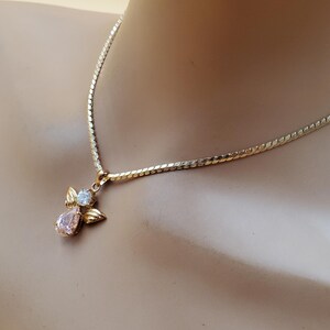 May include: A gold-coloured necklace with an angel pendant. The pendant has a pink, teardrop-shaped gemstone and a smaller, round, clear gemstone. The necklace is displayed on a mannequin.
