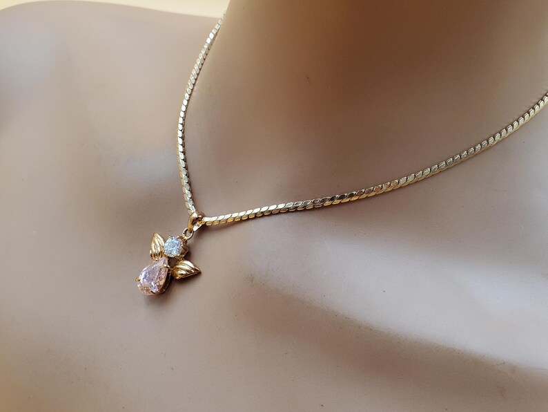 May include: A gold-coloured necklace with an angel pendant. The pendant has a pink, teardrop-shaped gemstone and a smaller, round, clear gemstone. The necklace is displayed on a mannequin.