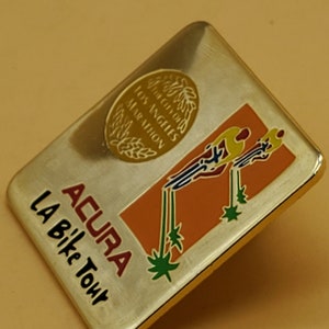 May include: Gold-colored metal pin with a red, orange, and green design. The pin features the text "ACURA LA Bike Tour" and a circular emblem with the text "The City of Los Angeles Marathon".