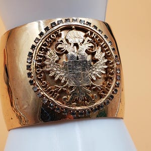 May include: Gold-tone cuff bracelet featuring a detailed emblem with an eagle and text. The wide band is embellished with small, dark-colored stones around the emblem. The bracelet has a polished finish and a vintage aesthetic.
