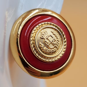 Puede incluir: Pendientes redondos dorados con centro rojo. El centro presenta un emblema dorado con corona, escudo y diseño de corona de laurel. Los pendientes tienen un acabado pulido, adecuado para ocasiones formales o cotidianas.