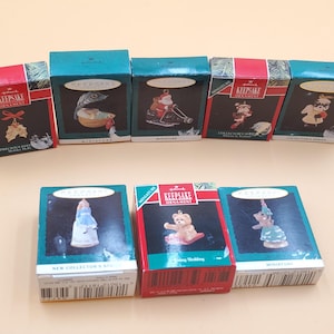 May include: A selection of Hallmark Keepsake miniature Christmas ornaments in their original boxes. The boxes are red and green, featuring various ornament designs such as a bear, angel, and other festive figures. The boxes are arranged on a light beige surface.
