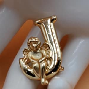 May include: A gold-colored brooch in the shape of the letter "J" with a small cherub figure. The cherub has wings and is seated on a crescent-shaped base. The brooch has a polished finish and is likely made of metal.