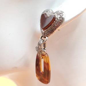 May include: An earring with a silver heart-shaped top and a rectangular amber-coloured pendant. The heart has a dark brown centre and ornate scrollwork. The pendant is a warm, translucent brown.
