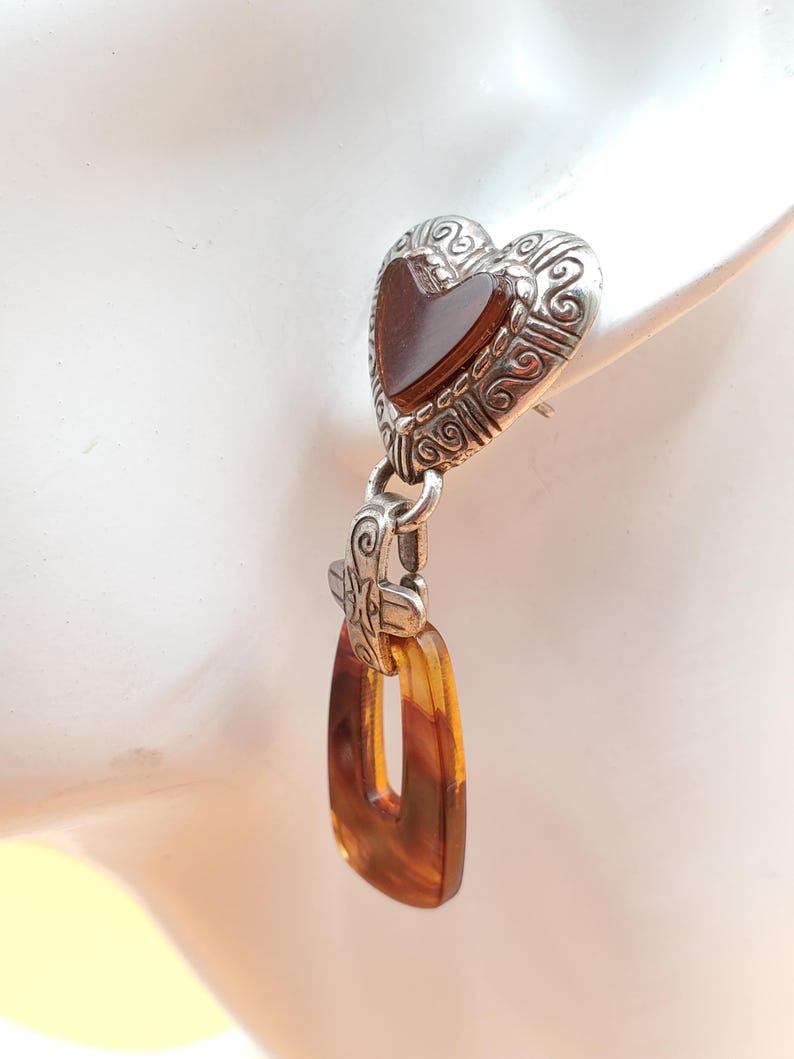 May include: An earring with a silver heart-shaped top and a rectangular amber-coloured pendant. The heart has a dark brown centre and ornate scrollwork. The pendant is a warm, translucent brown.