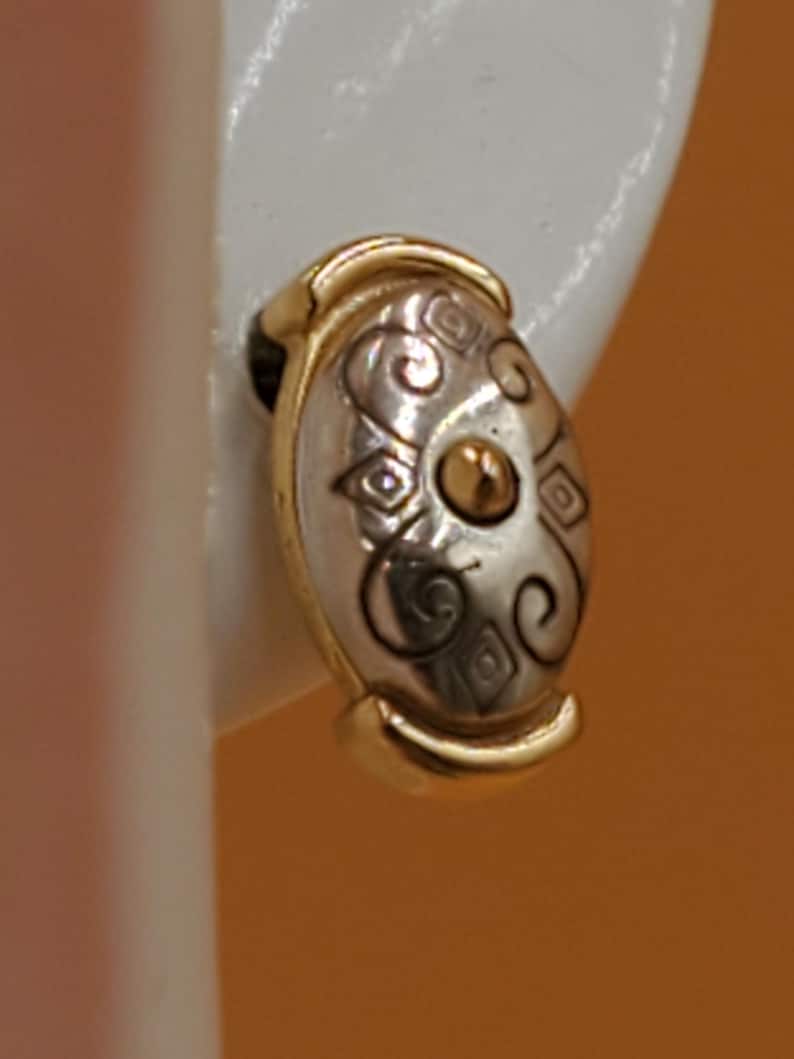 May include: Oval-shaped clip-on earring with a gold-tone frame and a silver-tone center. The center features a gold-tone dot and decorative swirls and diamond shapes. The earring is shown against a white background.