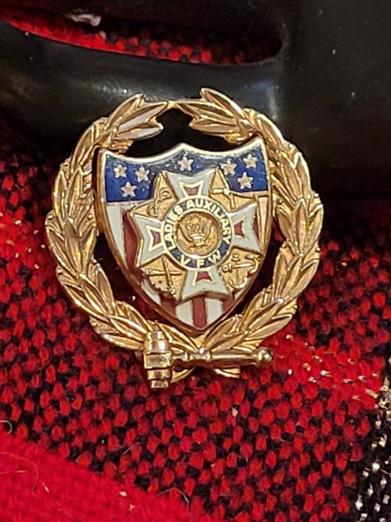 May include: Gold-toned pin with a wreath of leaves surrounding a shield with an American flag design. The shield also features a red, white, and blue cross with the words "Ladies Auxiliary VFW of the US" in a circle around it.