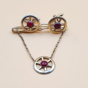 May include: A gold-toned tie bar and cufflinks set with a red stone in the centre of each wheel-shaped design.