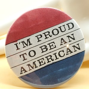 May include: A round button with a red, white and blue design. The button has the text "I'm Proud to Be An American" in black.