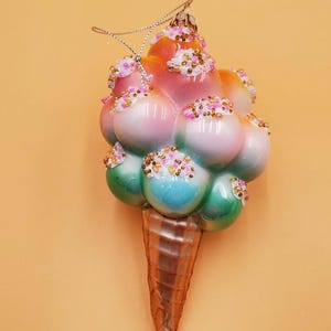 May include: A glass Christmas ornament shaped like an ice cream cone with pink, green, and blue ice cream and white sprinkles.